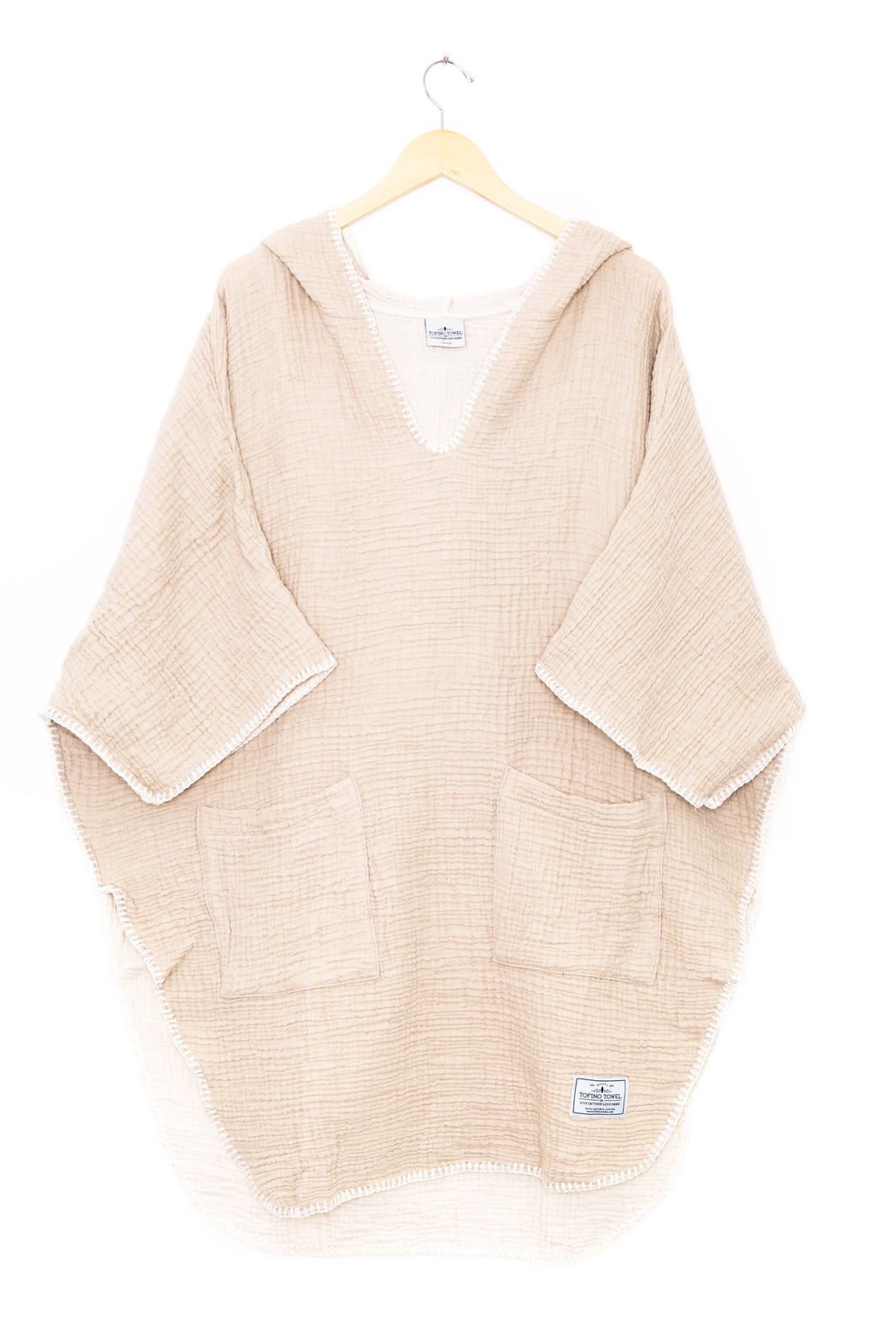 beige muslin poncho with curved hem and oversized hood and front pockets  on a wooden hanger against a white background #colour_beige