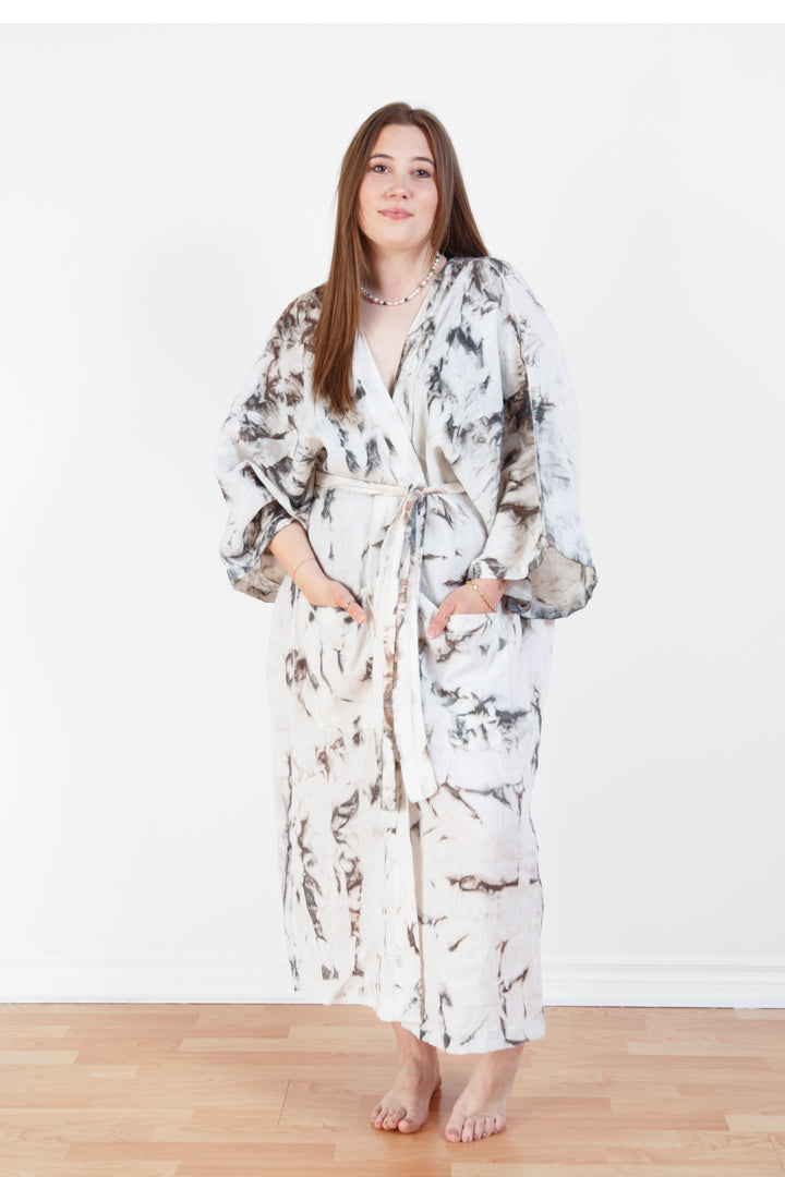 A woman standing in a room with a plain white wall, wearing a marble  tie-dyed linen belted cover-up robe with a belt around her waist.