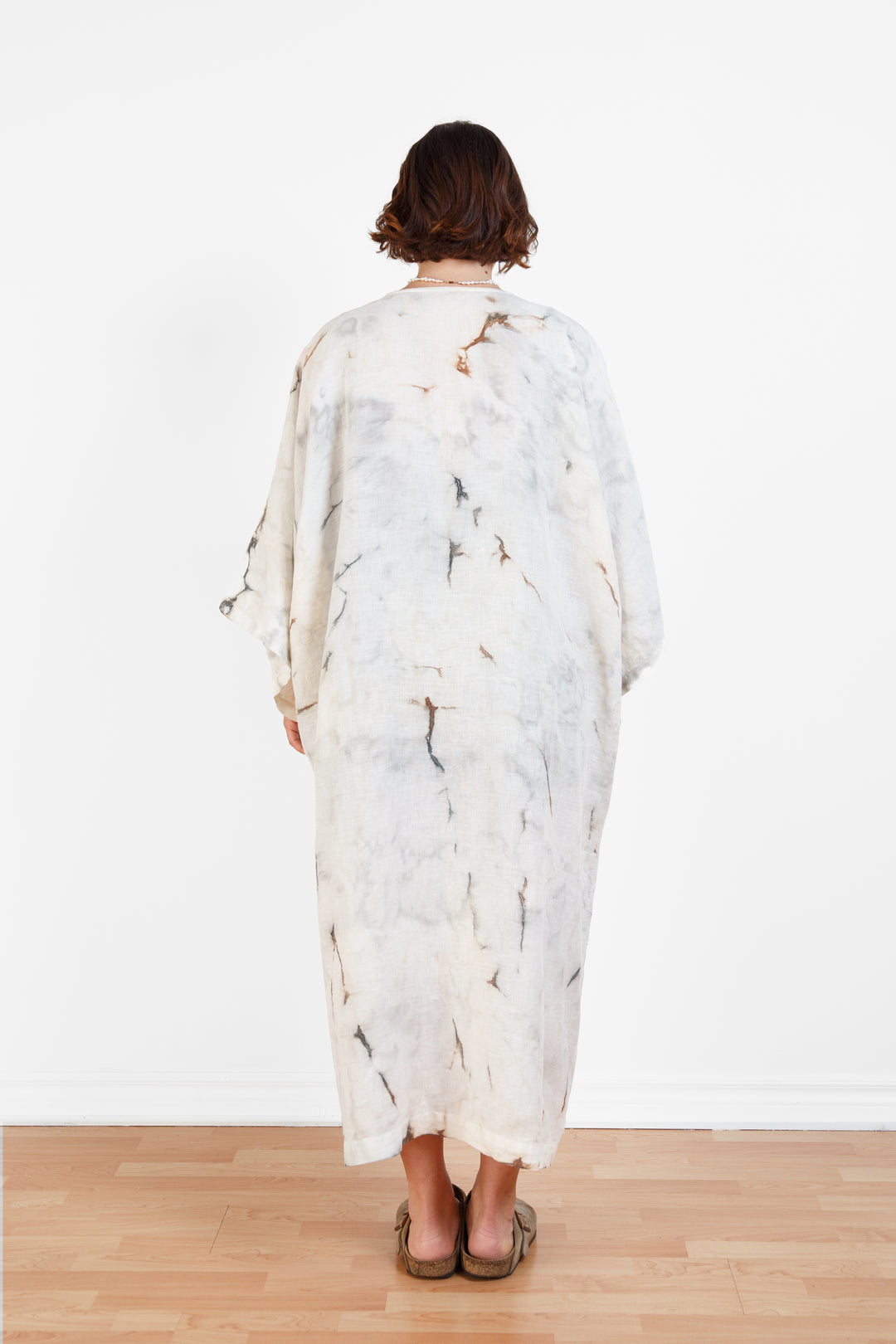 A woman standing backwards in a room with a plain white wall, wearing a marble  tie-dyed linen belted cover-up robe, open, untied