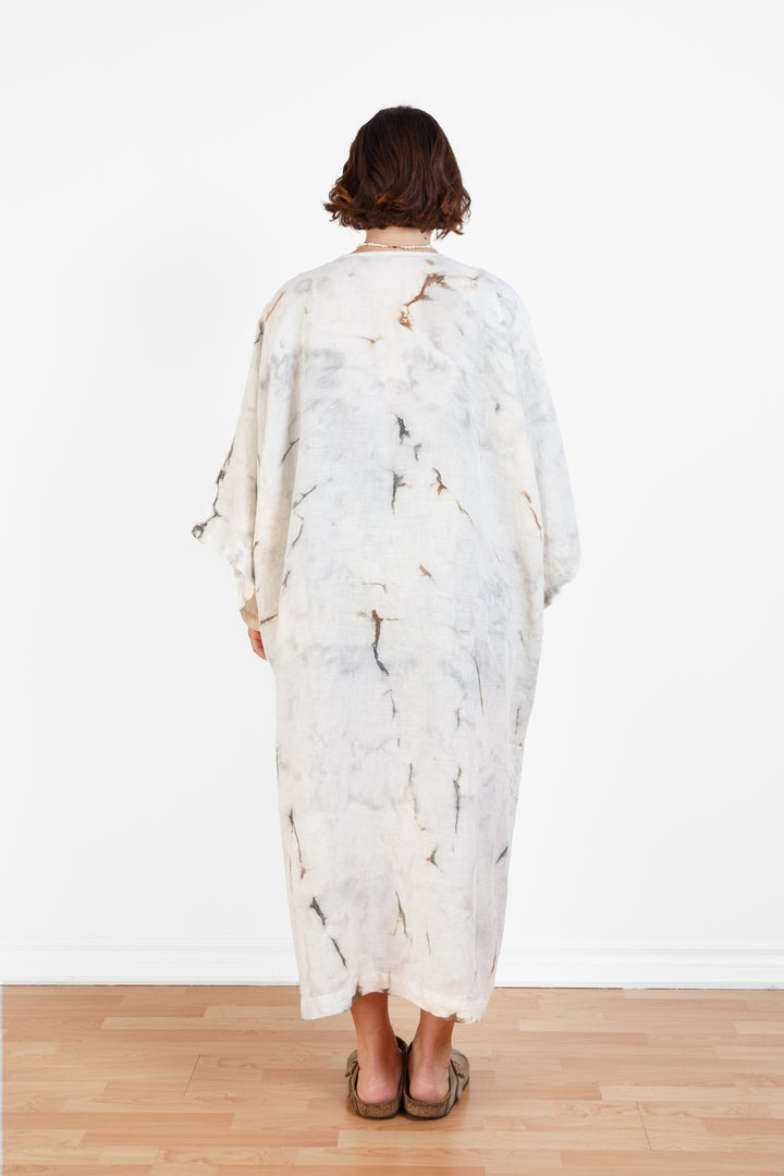 A woman standing backwards in a room with a plain white wall, wearing a marble  tie-dyed linen belted cover-up robe, open, untied
