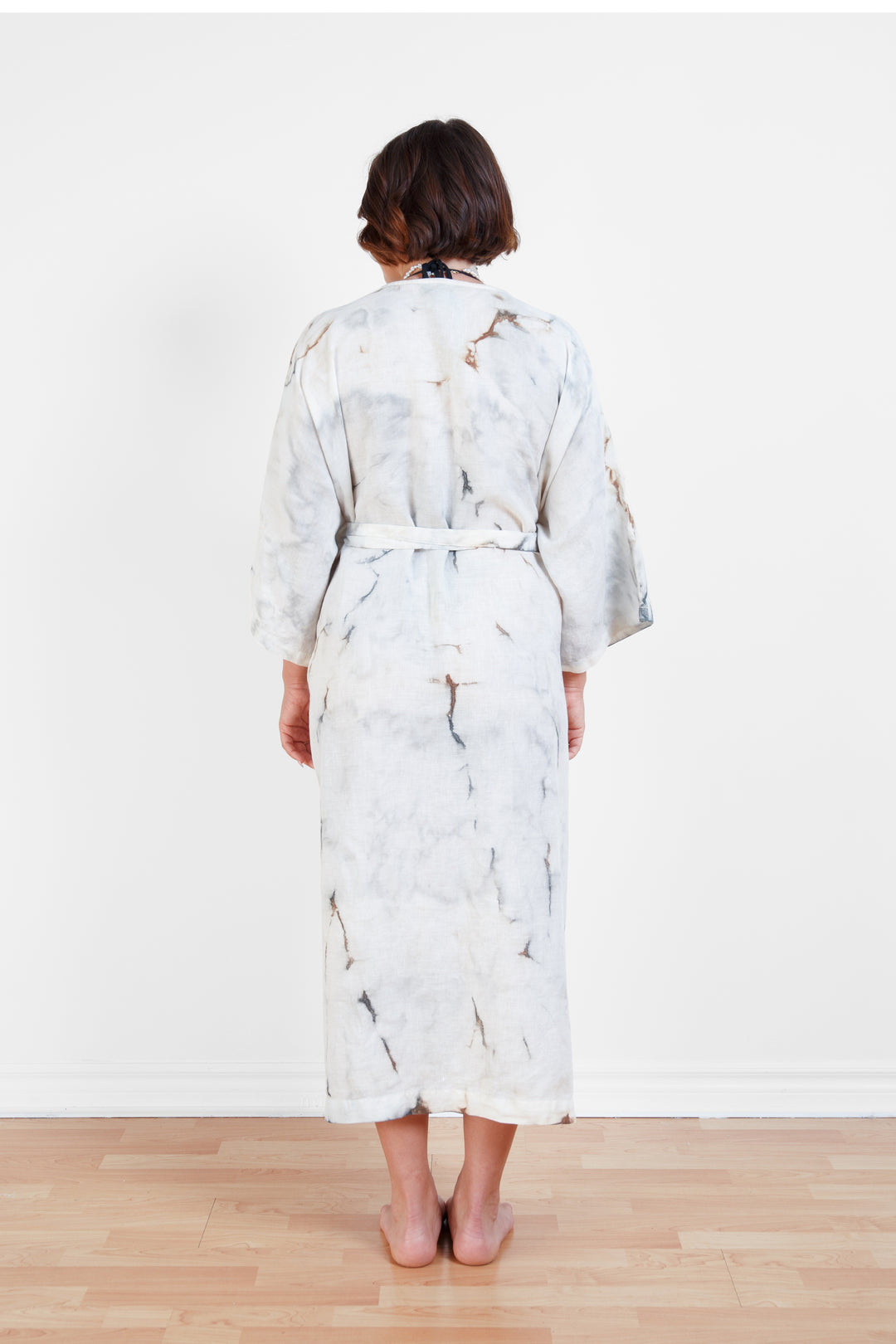 A woman standing backwards in a room with a plain white wall, wearing a marble  tie-dyed linen belted cover-up robe, tied