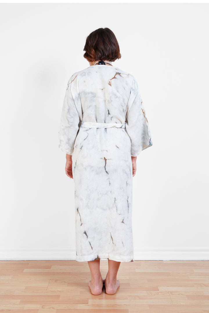 A woman standing backwards in a room with a plain white wall, wearing a marble  tie-dyed linen belted cover-up robe, tied