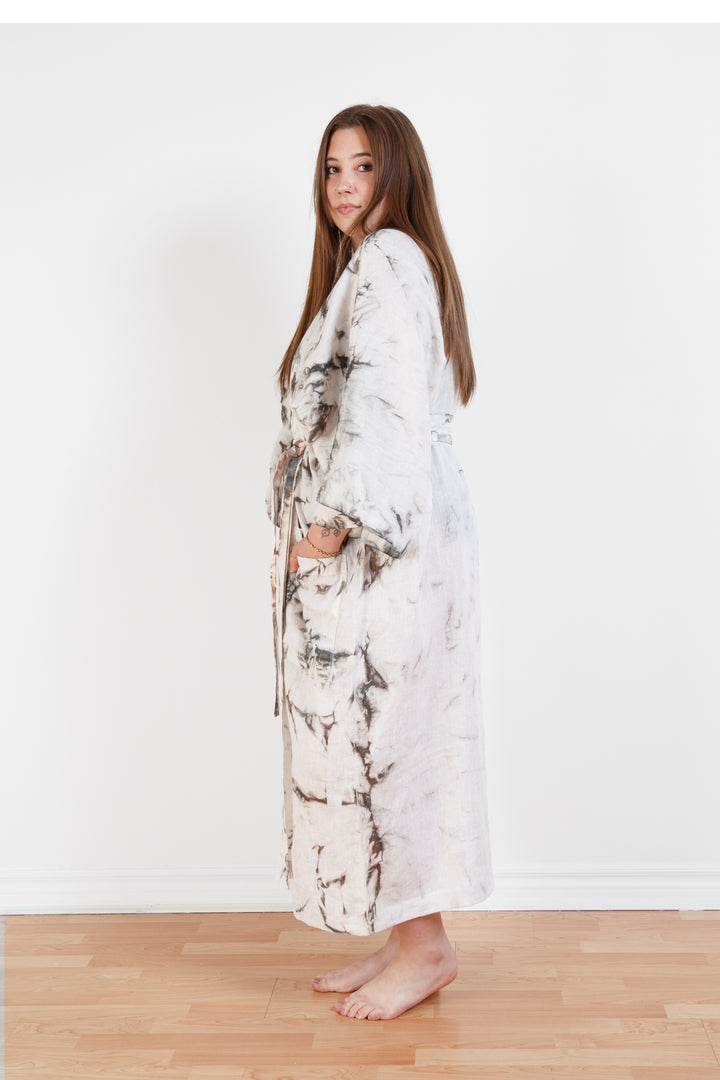 A woman standing in a room with a plain white wall, wearing a marble  tie-dyed linen belted cover-up robe