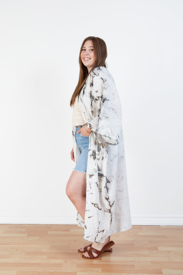 A woman standing in a room with a plain white wall, wearing a marble  tie-dyed linen cover-up robe, open