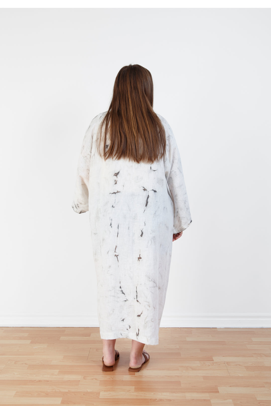 A woman standing backwards in a room with a plain white wall, wearing a marble  tie-dyed linen belted cover-up robe, untied