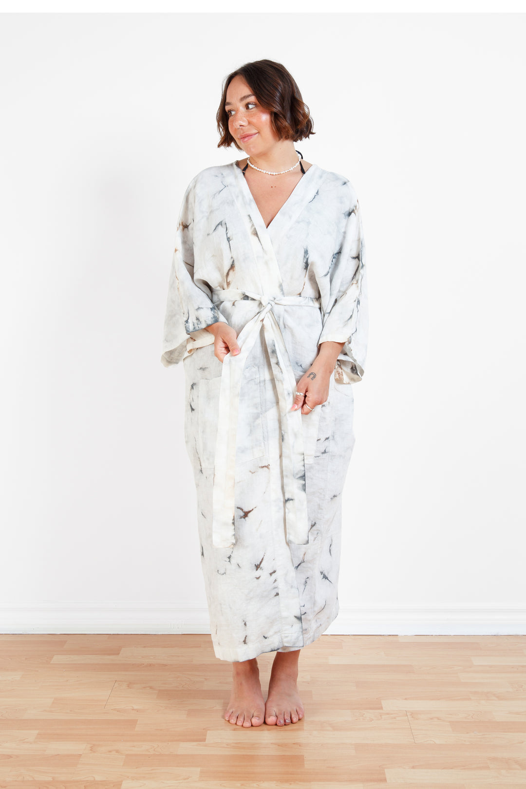 A woman standing in a room with a plain white wall, wearing a marble tie-dyed linen belted cover-up robe, tied