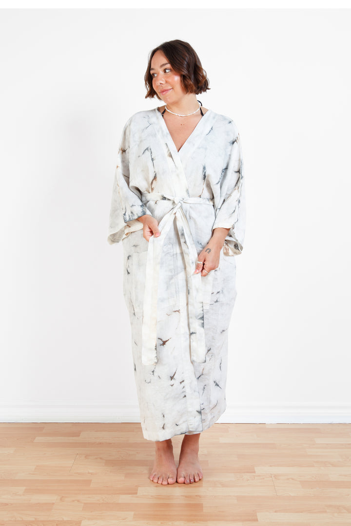 A woman standing in a room with a plain white wall, wearing a marble tie-dyed linen belted cover-up robe, tied