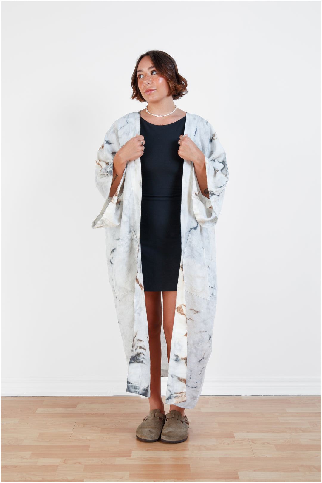 A woman standing in a room with a plain white wall, wearing a marble  tie-dyed linen belted cover-up robe, open, untied