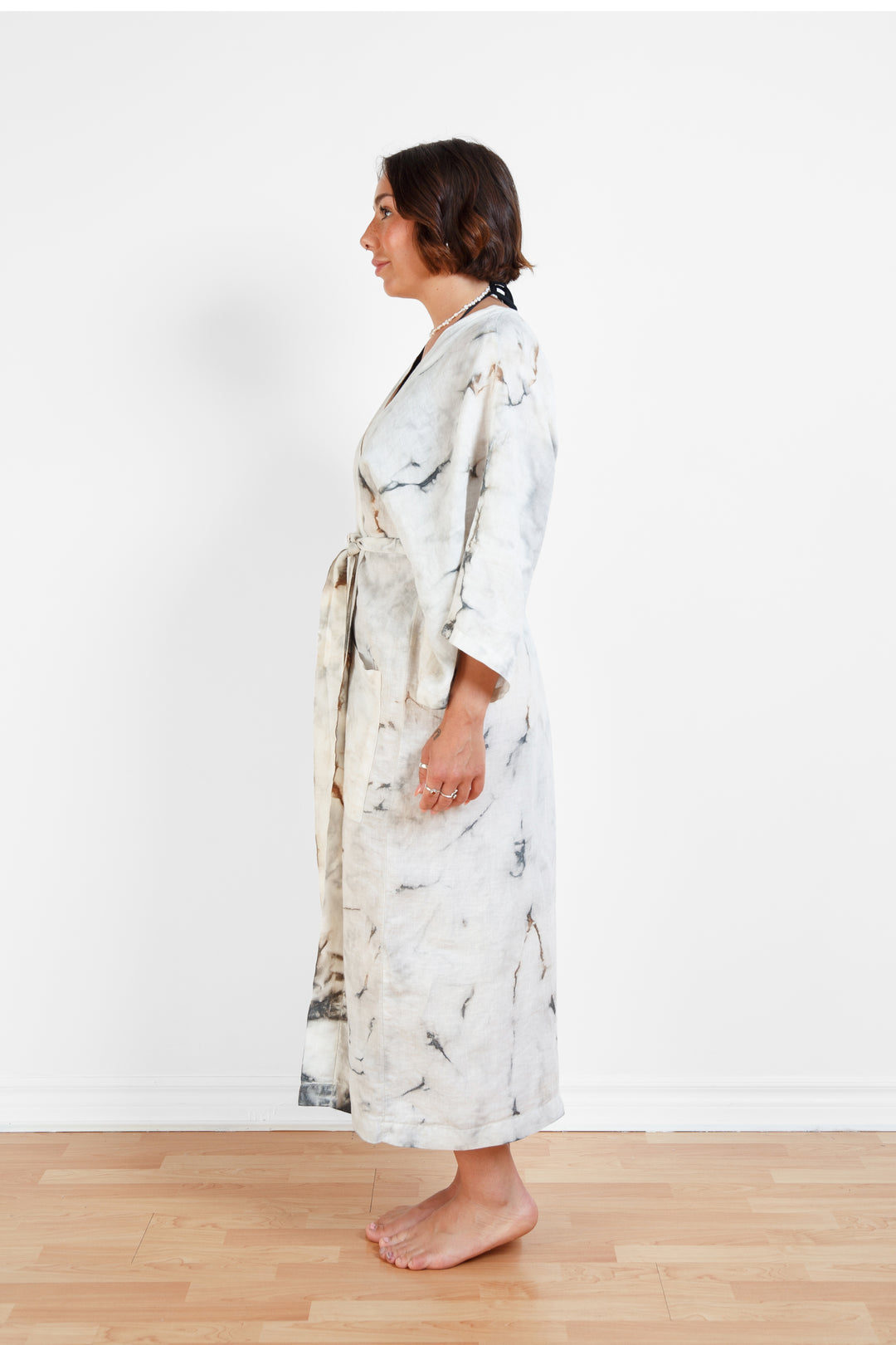 A woman standing in a room with a plain white wall, wearing a marble  tie-dyed linen belted cover-up robe, tied