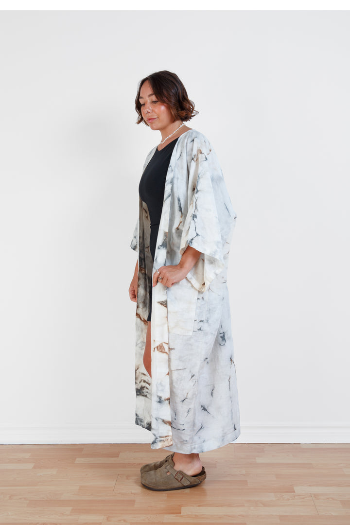 A woman standing in a room with a plain white wall, wearing a marble  tie-dyed linen belted cover-up robe, open, untied
