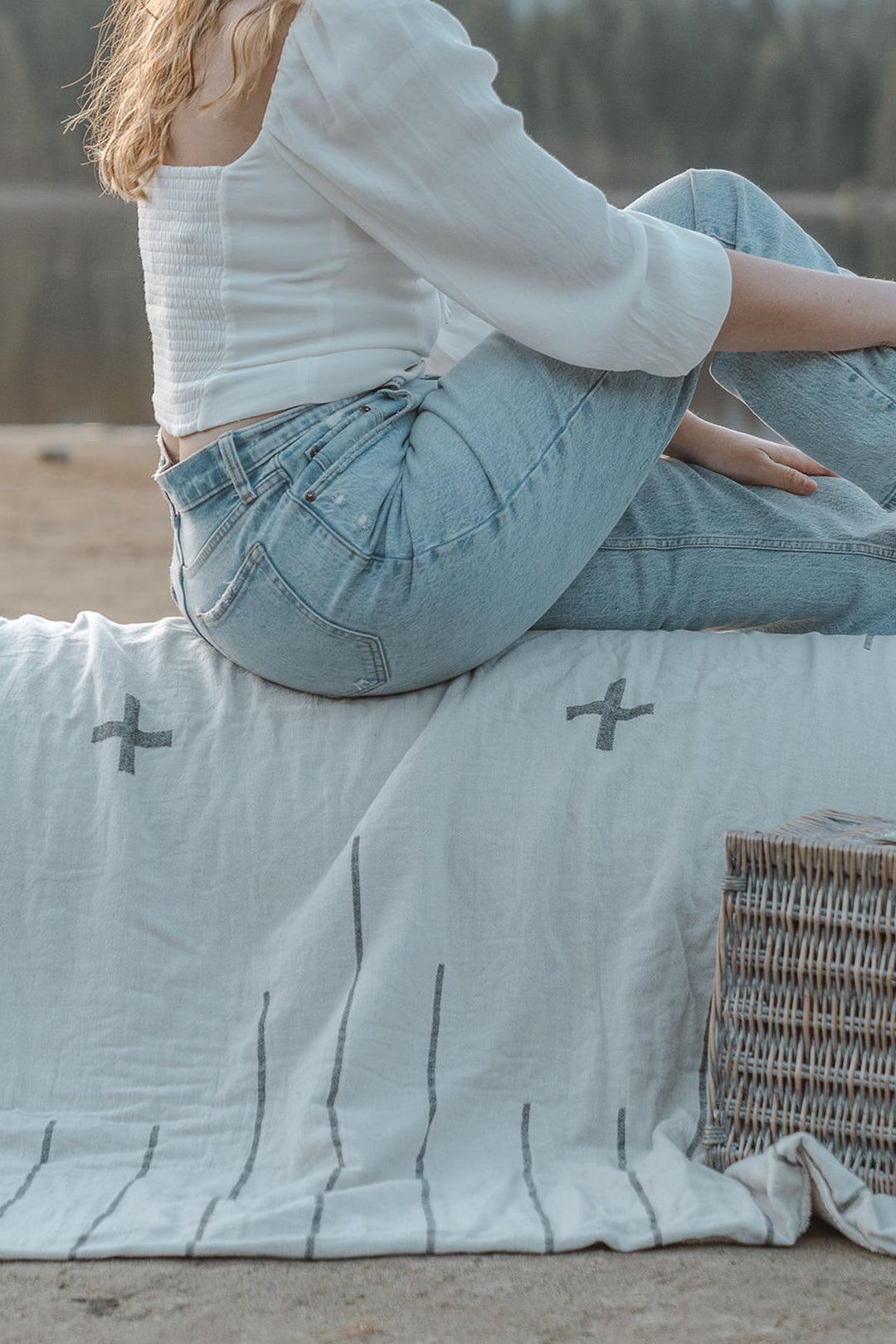 girl with blonde hair wearing a white blouse and faded jeans sitting on an off-white blanket with simple minimal dark grey design outdoors on a beach #colour_whitecap