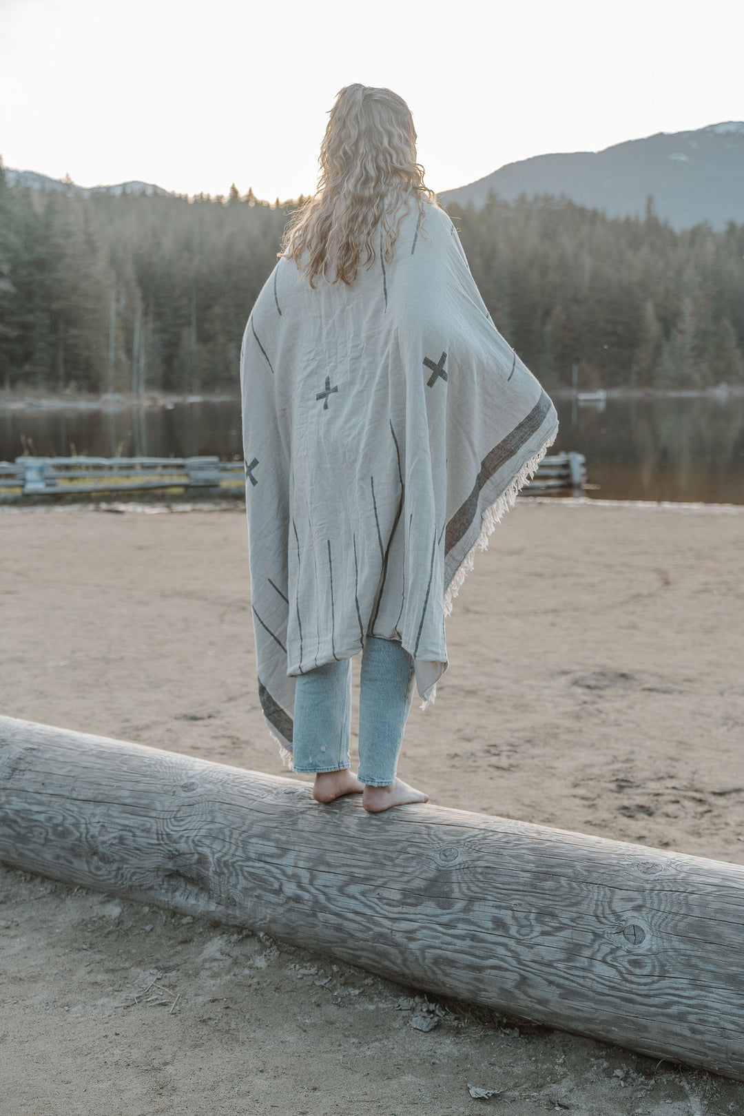 girl with blond hair wearing light blue jeans standing on a log on a sandy beach wrapped in an off white blanket with a simple minimal design in dark grey and eye lash fringe lake and mountains in the background #colour_whitecap