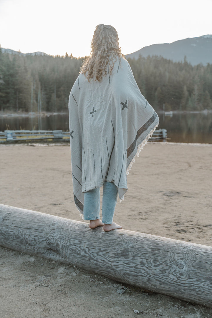 girl with blond hair wearing light blue jeans standing on a log on a sandy beach wrapped in an off white blanket with a simple minimal design in dark grey and eye lash fringe lake and mountains in the background #colour_whitecap