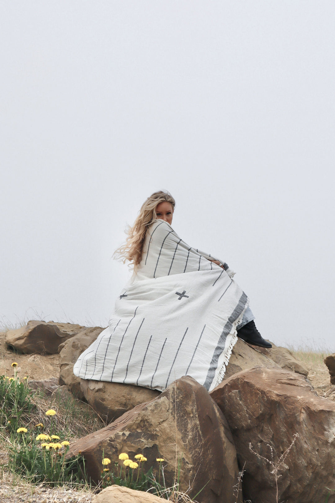 girl with blonde hair sitting on rocks 
 wrapped in an off white blanket with a simple dark grey pattern overcast sky in the background and dandillions in foreground  #colour_whitecap