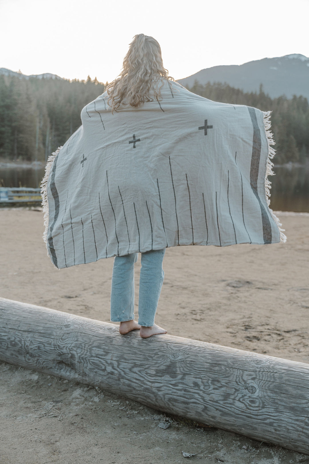 girl with blond hair wearing light blue jeans standing on a log on a sandy beach wrapped in an off white blanket with a simple minimal design in dark grey and eye lash fringe lake and mountains in the background #colour_whitecap