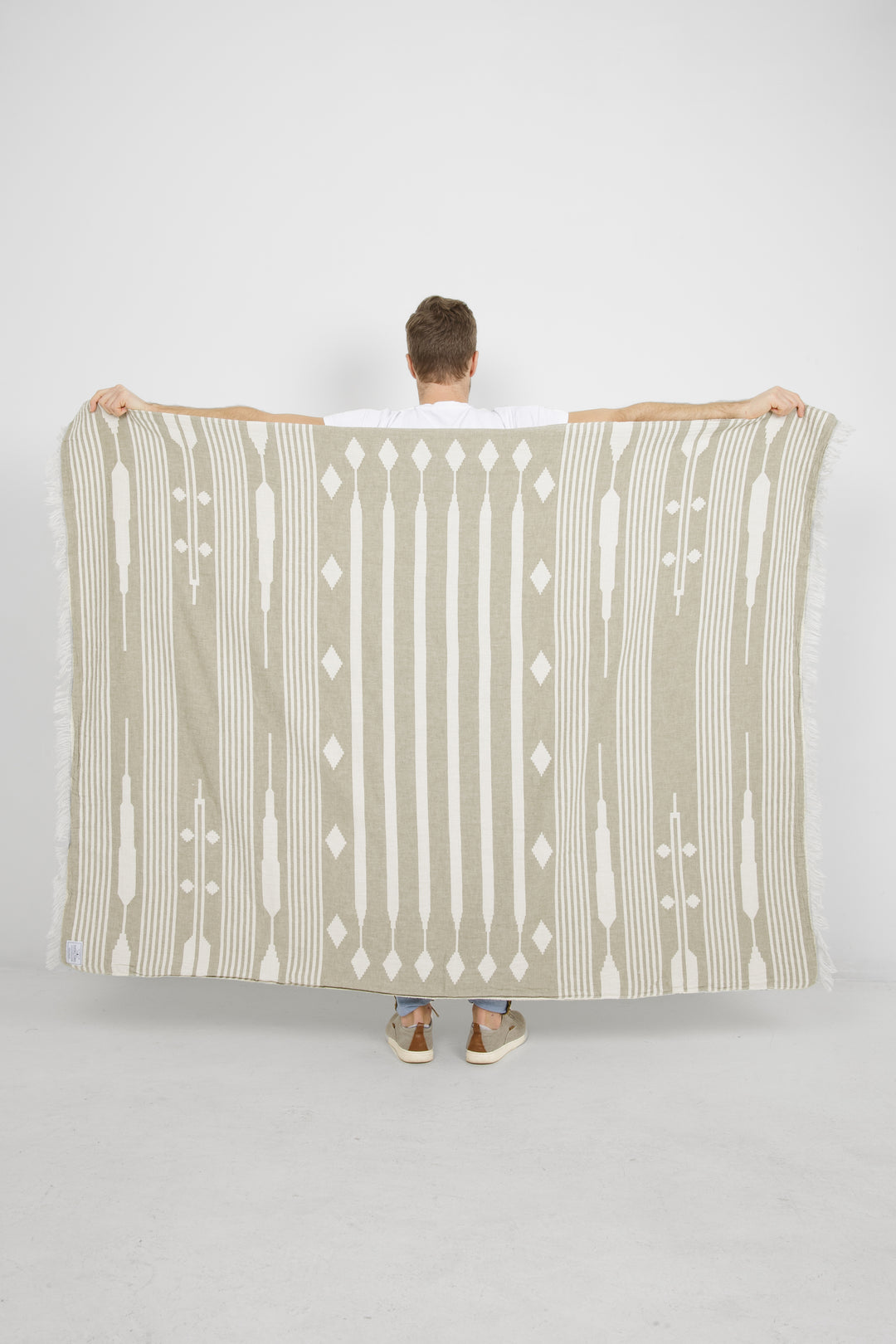 male standing backwards spreading a khaki and cream traditional geometric, fleece lined throw with eyelash fringe behind him #colour_khaki