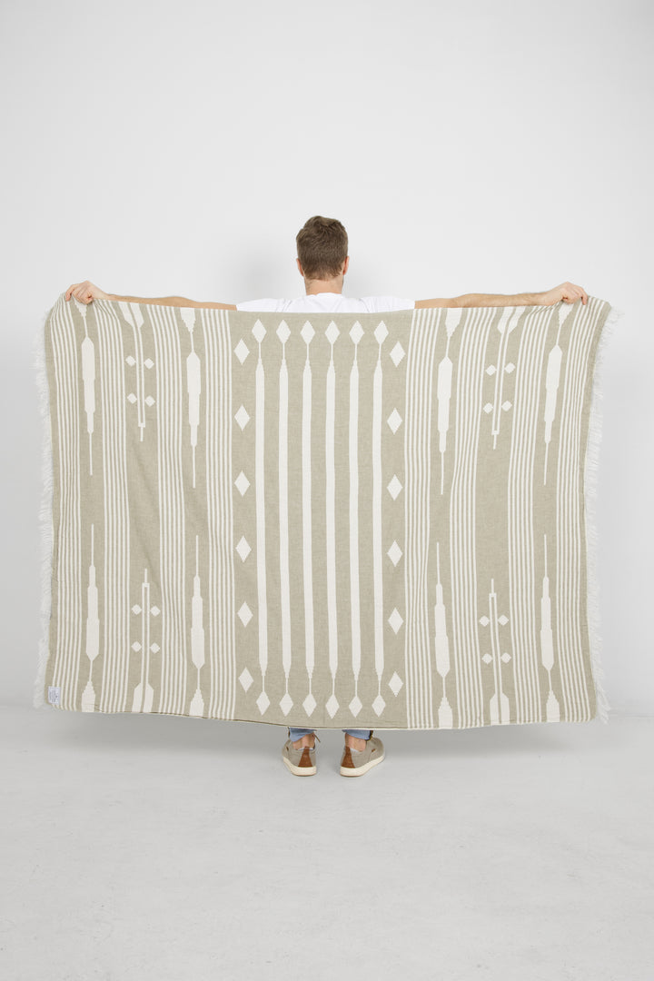 male standing backwards spreading a khaki and cream traditional geometric, fleece lined throw with eyelash fringe behind him #colour_khaki