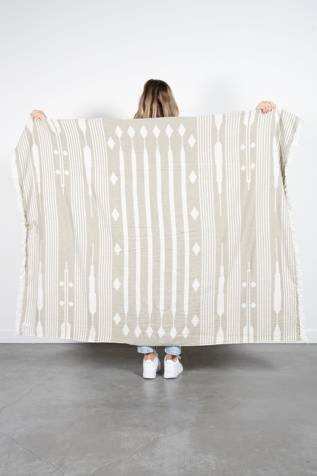 woman standing backwards spreading a khaki and cream traditional geometric, fleece lined throw with eyelash fringe behind her #colour_khaki