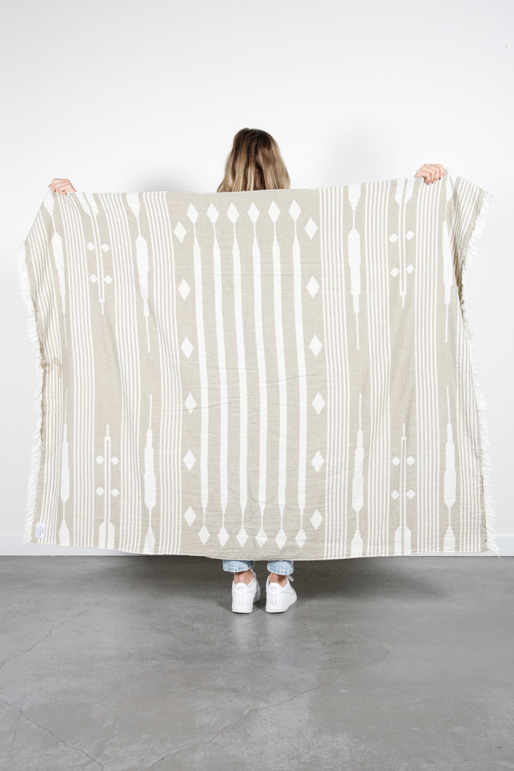 woman standing backwards spreading a khaki and cream traditional geometric, fleece lined throw with eyelash fringe behind her #colour_khaki