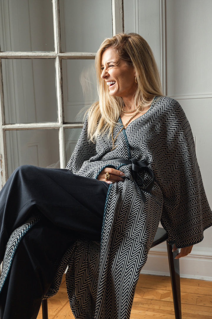 Woman sitting wearing a long, herringbone patterned cardigan with blue whipstitch and pockets over black pants