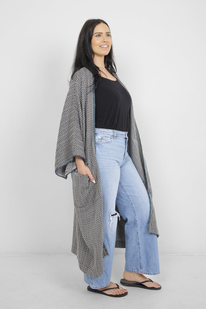 Woman wearing a long, herringbone patterned cardigan with blue whipstitch and pockets over a black top and blue jeans on a white background