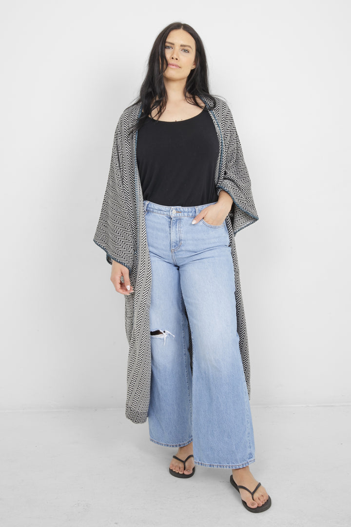 Woman wearing a long, herringbone patterned cardigan with blue whipstitch and pockets over a black top and blue jeans on a white background