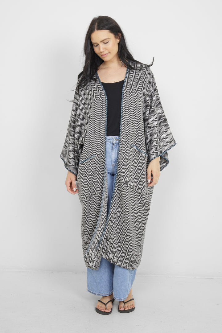 Woman wearing a long, herringbone patterned cardigan with blue whipstitch and pockets over a black top and blue jeans on a white background