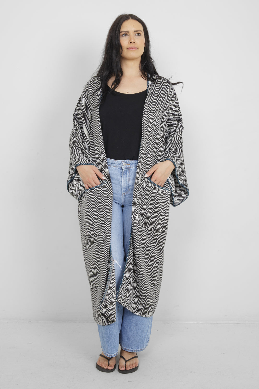 Woman wearing a long, herringbone patterned cardigan with blue whipstitch and pockets over a black top and blue jeans on a white background