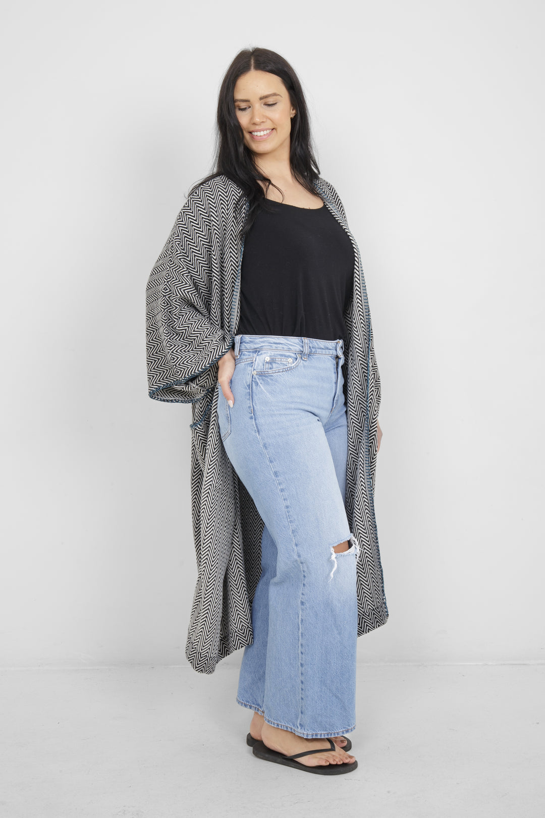 Woman wearing a long, herringbone patterned cardigan with blue whipstitch and pockets over a black top and blue jeans on a white background
