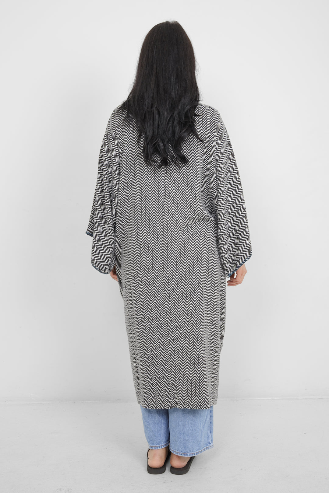Woman standing backwards wearing a long, herringbone patterned cardigan with blue whipstitch over blue jeans on a white background