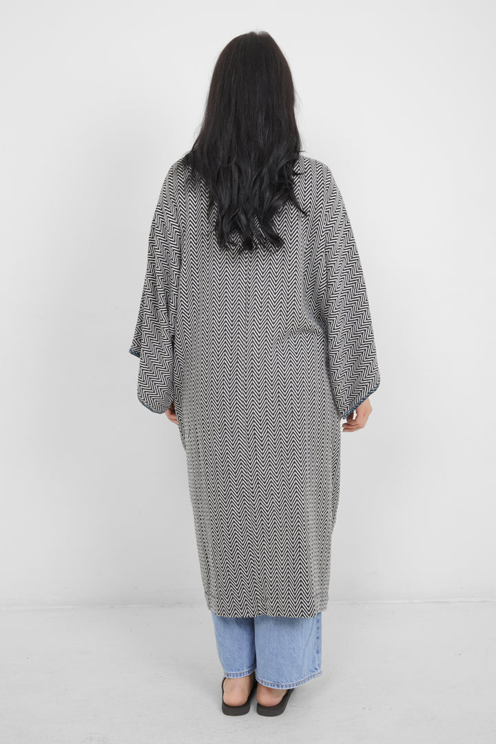 Woman standing backwards wearing a long, herringbone patterned cardigan with blue whipstitch over blue jeans on a white background