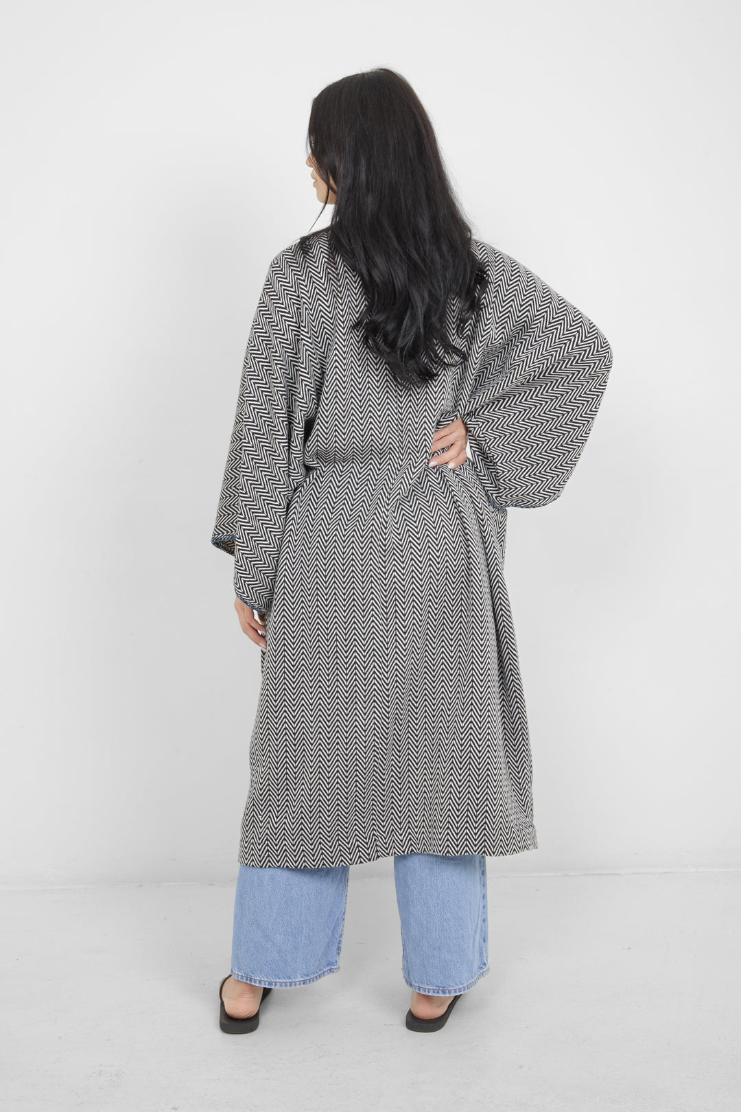 Woman standing backwards wearing a long, herringbone patterned cardigan with blue whipstitch over blue jeans on a white background