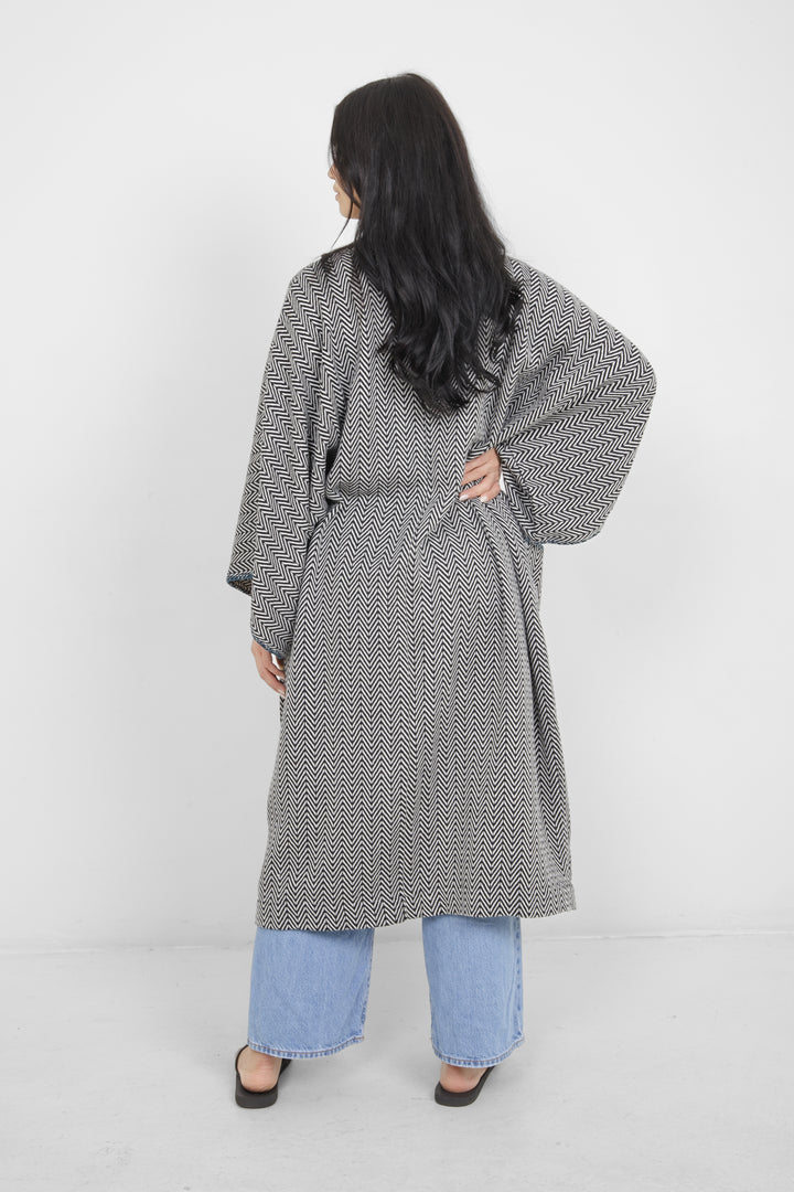 Woman standing backwards wearing a long, herringbone patterned cardigan with blue whipstitch over blue jeans on a white background