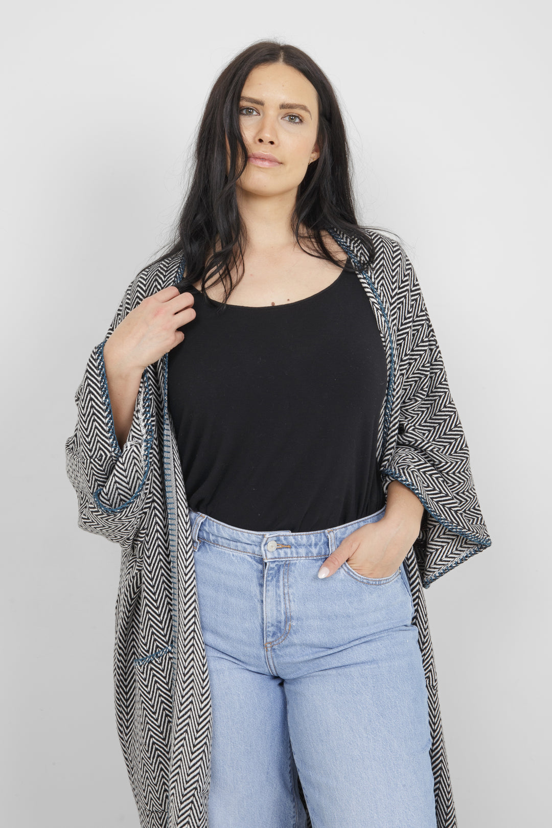 Woman wearing a long, herringbone patterned cardigan with blue whipstitch and pockets over a black top and blue jeans on a white background