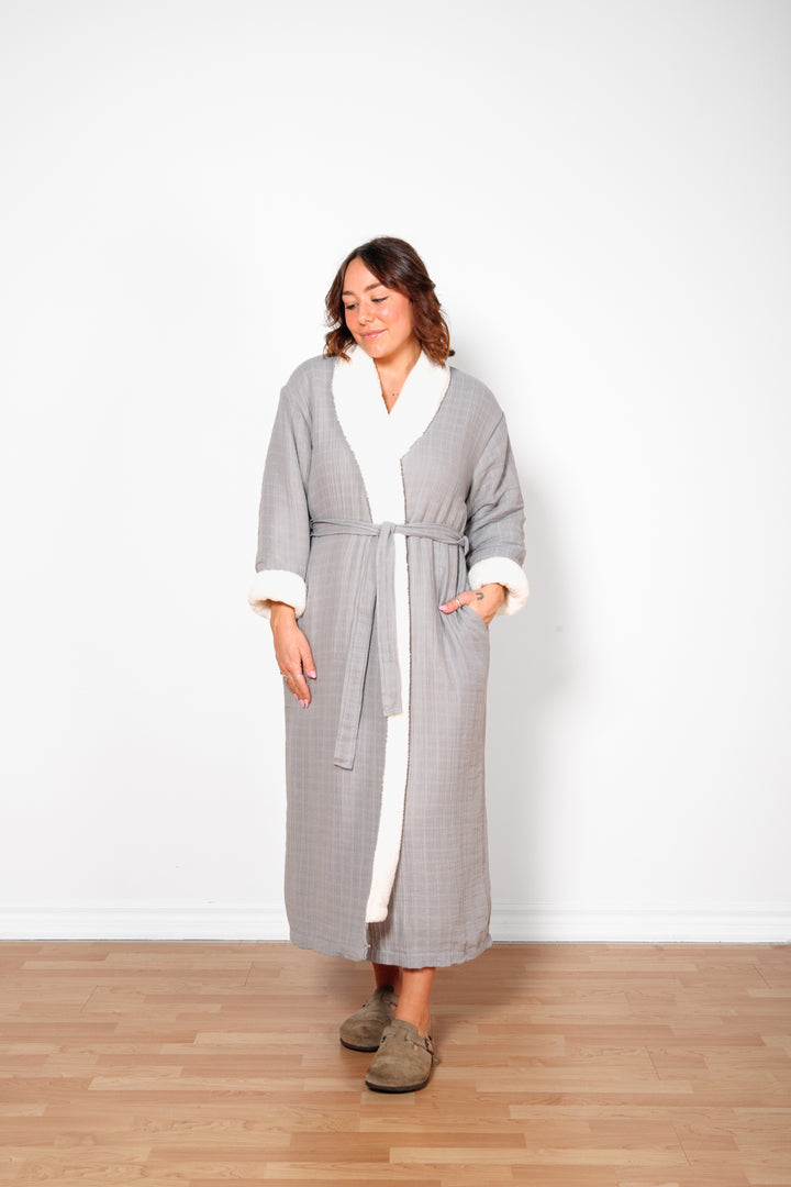 A woman wearing a long grey belted fleece lined muslin robe with pockets. #colour_grey