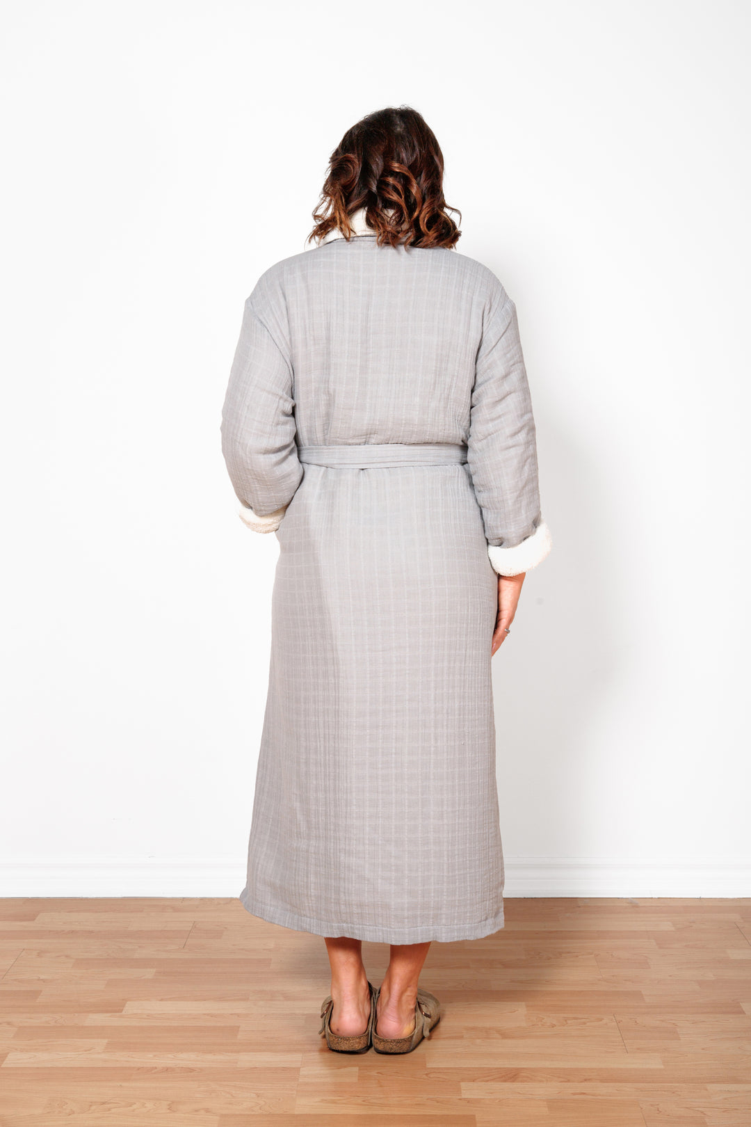 A woman standing backwards wearing a long grey belted fleece lined muslin robe with pockets. #colour_grey