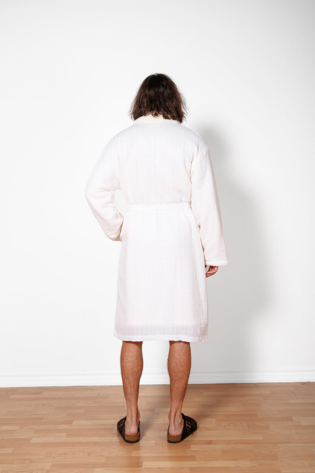 a man standing backwards wearing long white belted fleece lined muslin robe with pockets. #colour_white
