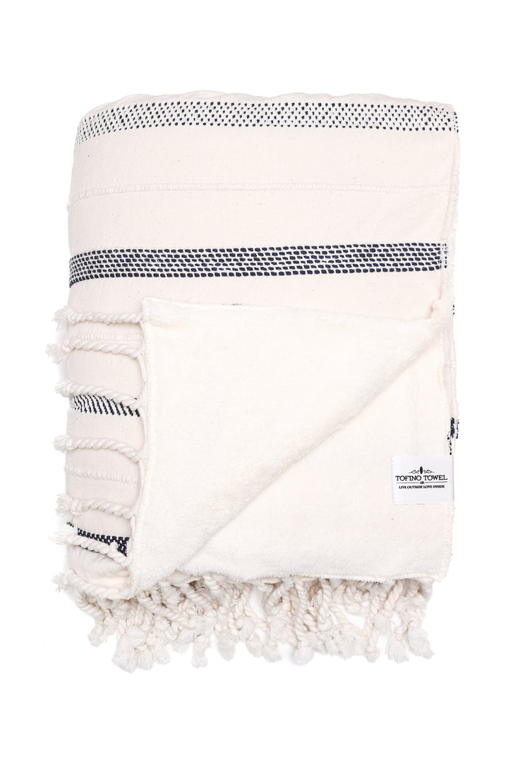 an off white fleece lined throw with different shades of navy stripes and tassels folded with one corner turned back on a white background