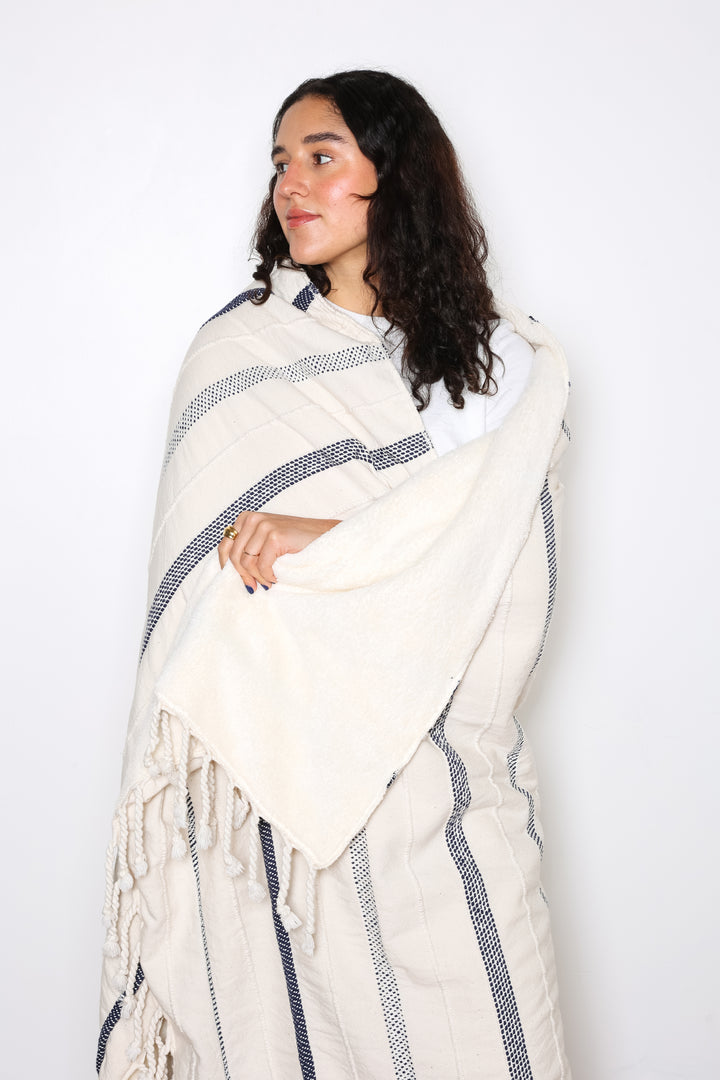 a women wrapped in an off white fleece lined throw with different shades of navy stripes and tassels