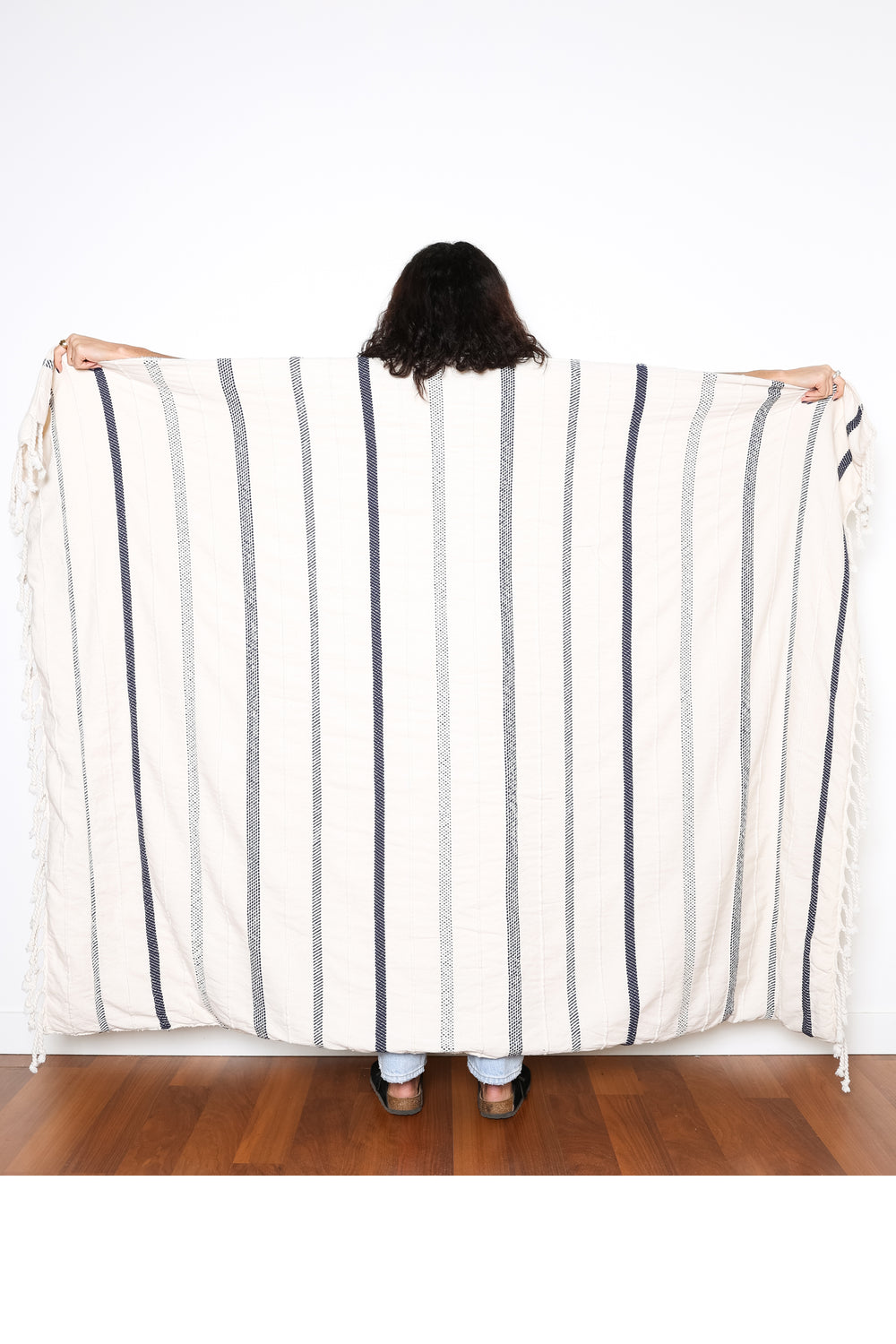 a women standing backwards holding out an off white fleece lined throw with different shades of navy stripes and tassels behind her