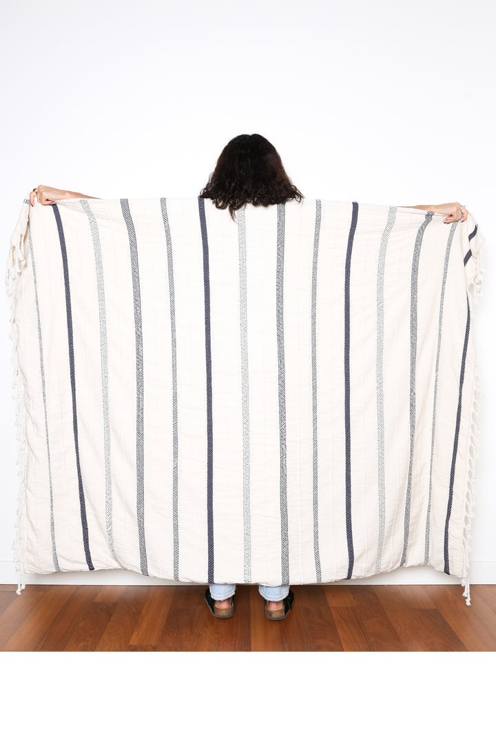 a women standing backwards holding out an off white fleece lined throw with different shades of navy stripes and tassels behind her