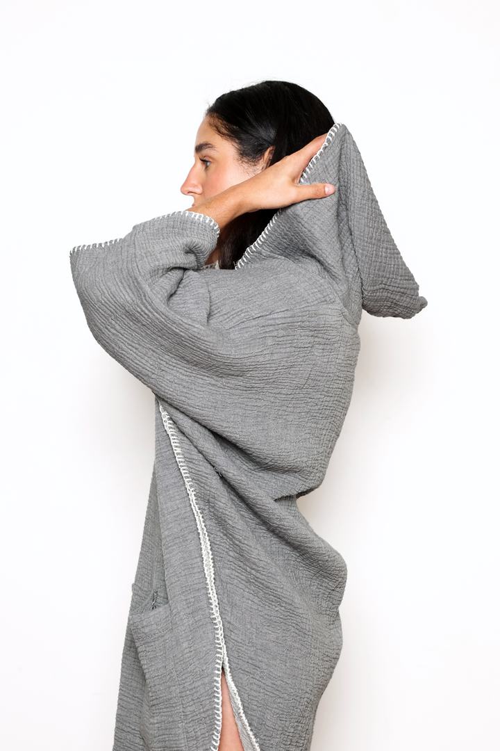 woman turned sideways wearing a  dark grey muslin poncho with curved hem and oversized hood and front pockets against a white background #colour_grey