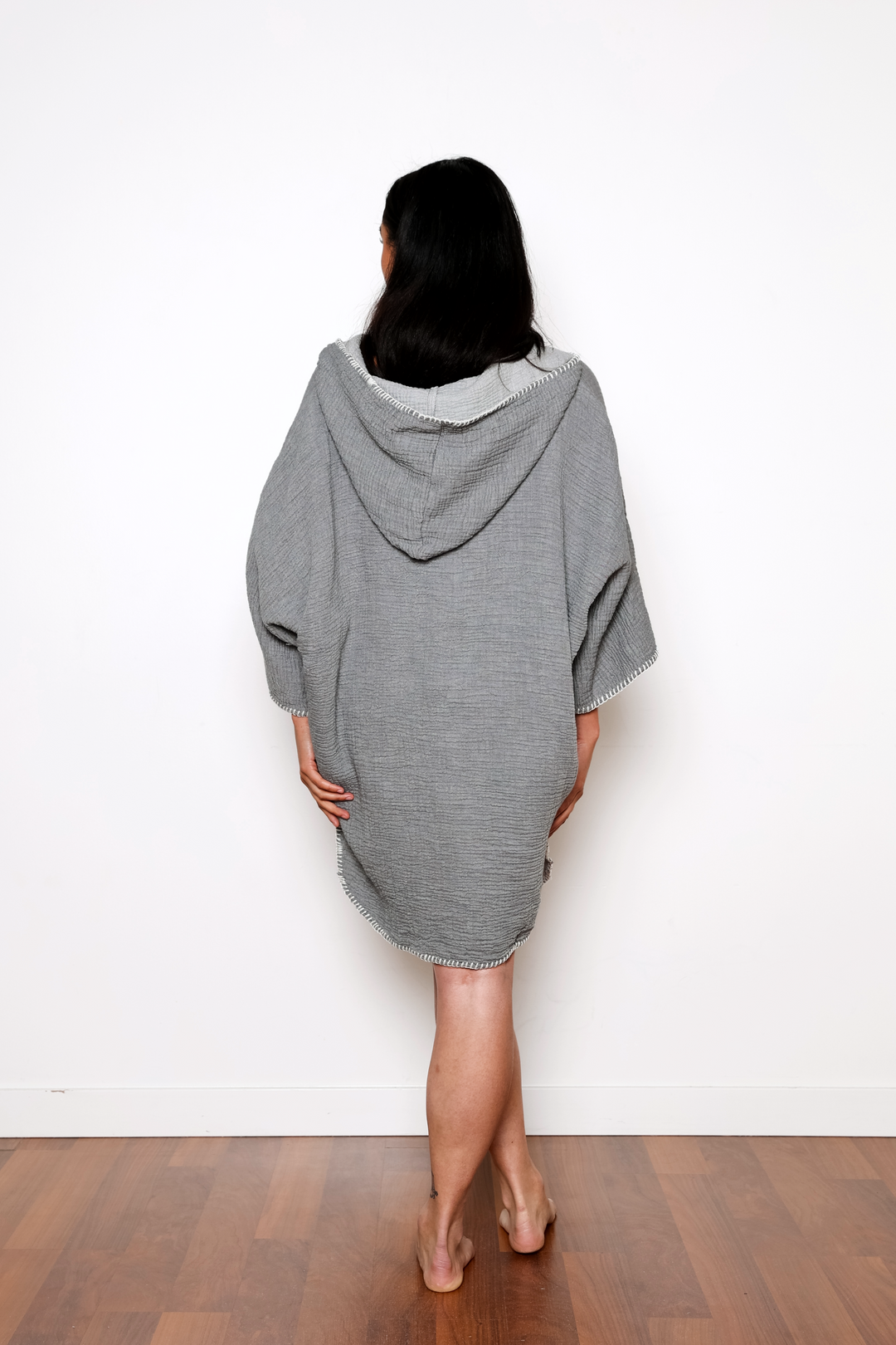 woman standing backwards wearing a  dark grey muslin poncho with curved hem and oversized hood and front pockets against a white background #colour_grey