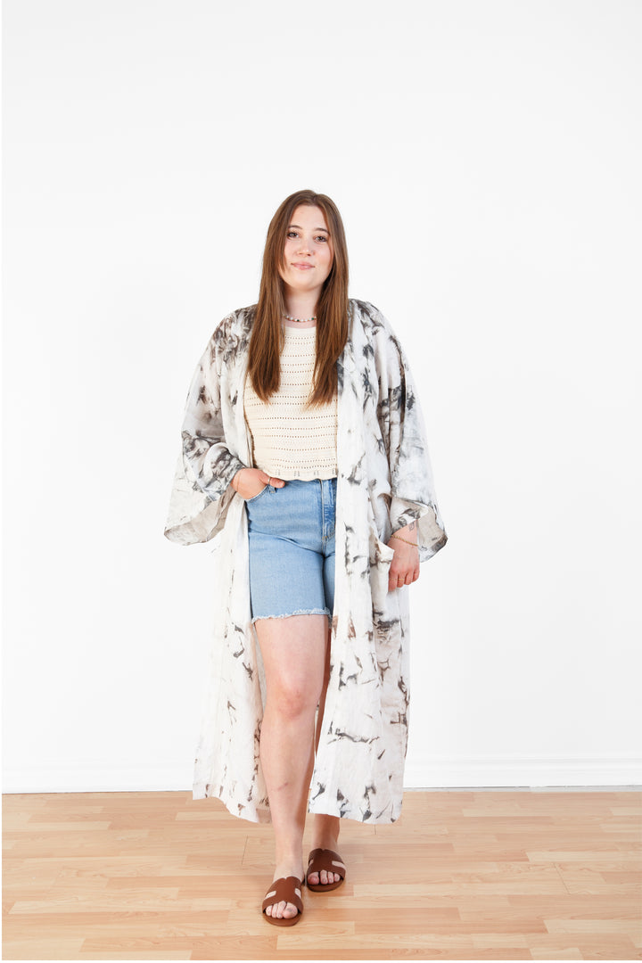 A woman standing in a room with a plain white wall, wearing a marble tie-dyed linen belted cover-up robe open