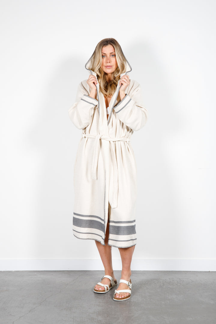 A woman standing wearing a medium length beige hooded belted terry robe with gray stripes, and pockets. 