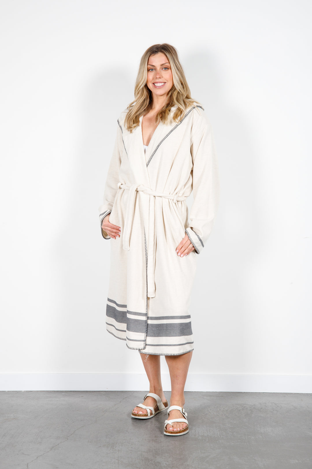 A woman standing wearing a medium length beige hooded belted terry robe with gray stripes, and pockets. 