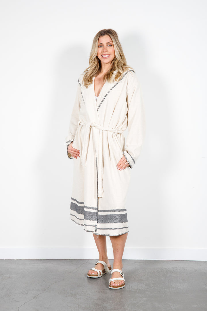 A woman standing wearing a medium length beige hooded belted terry robe with gray stripes, and pockets. 