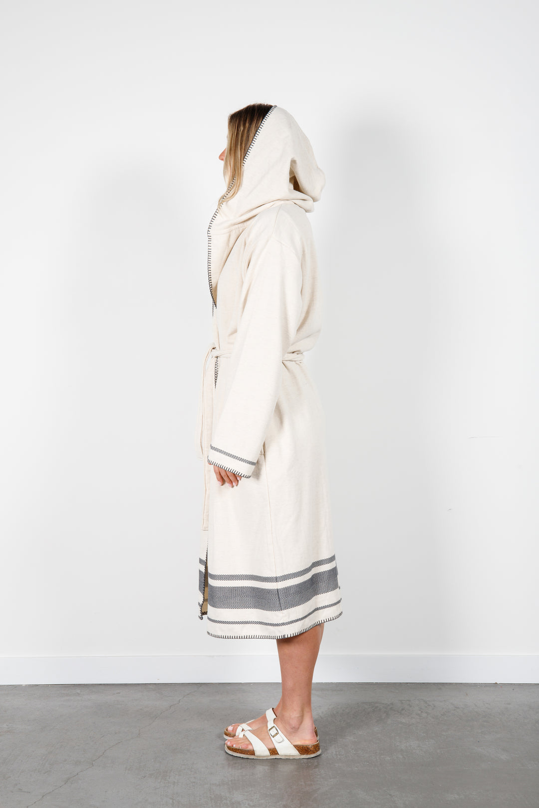 A woman standing wearing a medium length beige hooded belted terry robe with gray stripes, and pockets. Hood is up.