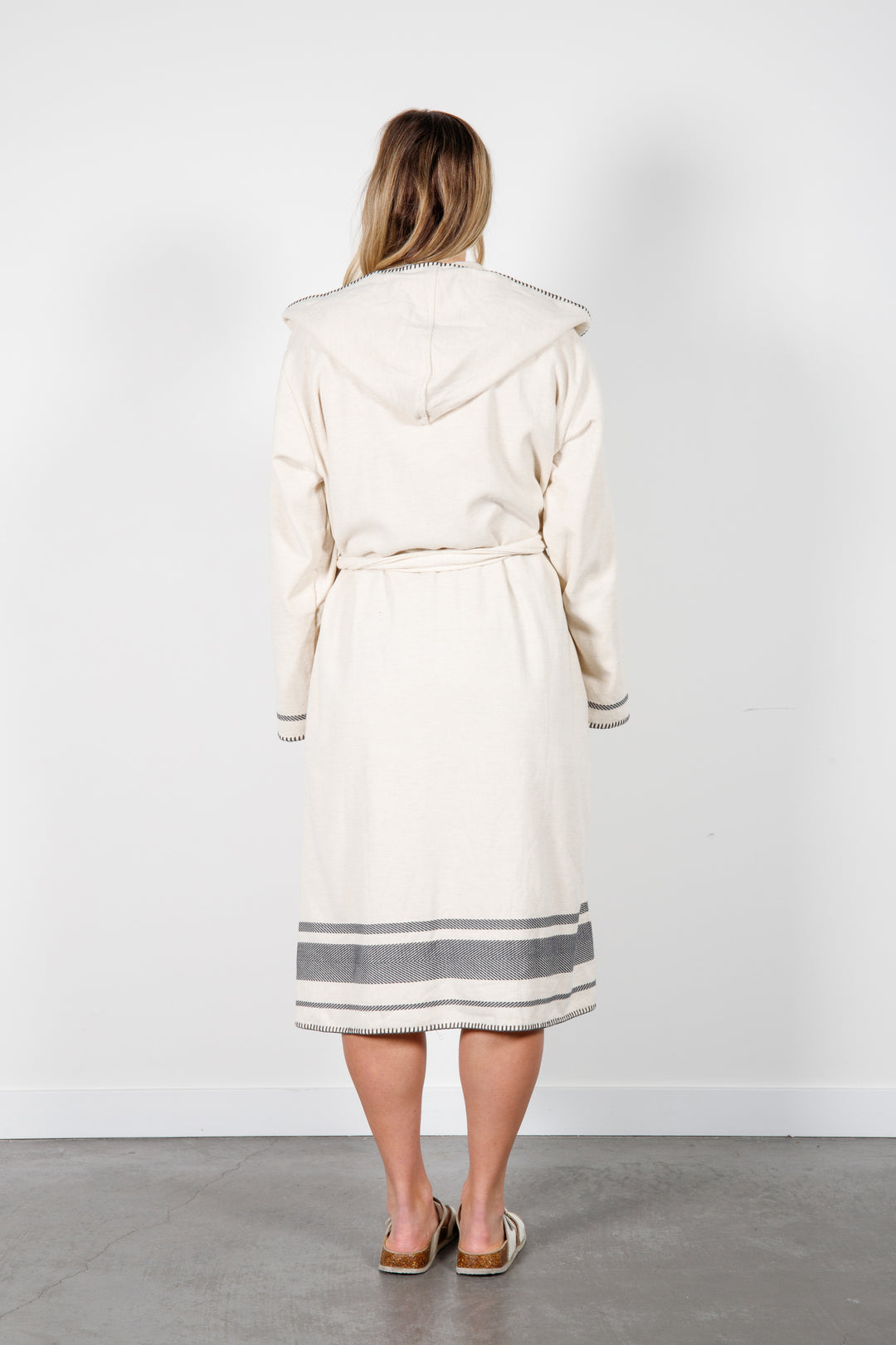 A woman standing backwards wearing a medium length beige hooded belted terry robe with gray stripes, and pockets. 