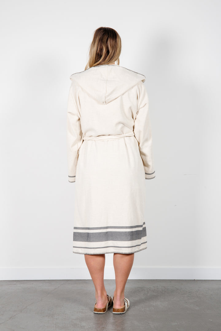A woman standing backwards wearing a medium length beige hooded belted terry robe with gray stripes, and pockets. 
